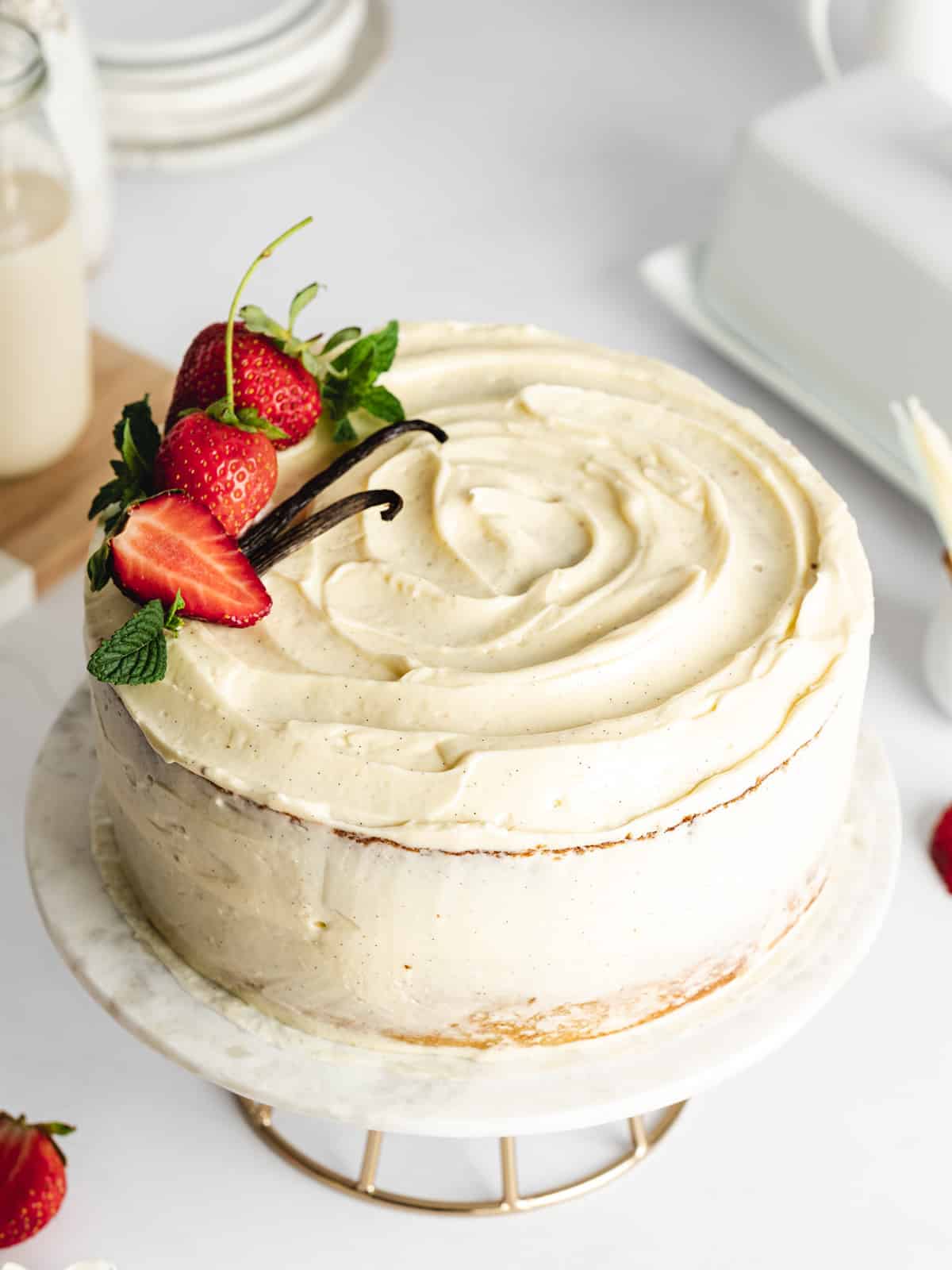 a vanilla cake on a marble cake stand with fresh strawberries, mint leaves, and vanilla pods on top.