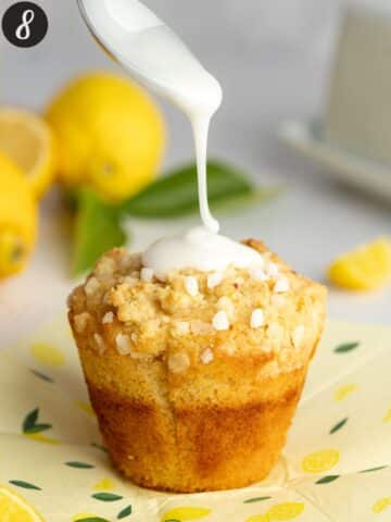 a vegan lemon muffin on lemon parchment paper with lemon glaze being drizzled over it with a spoon.