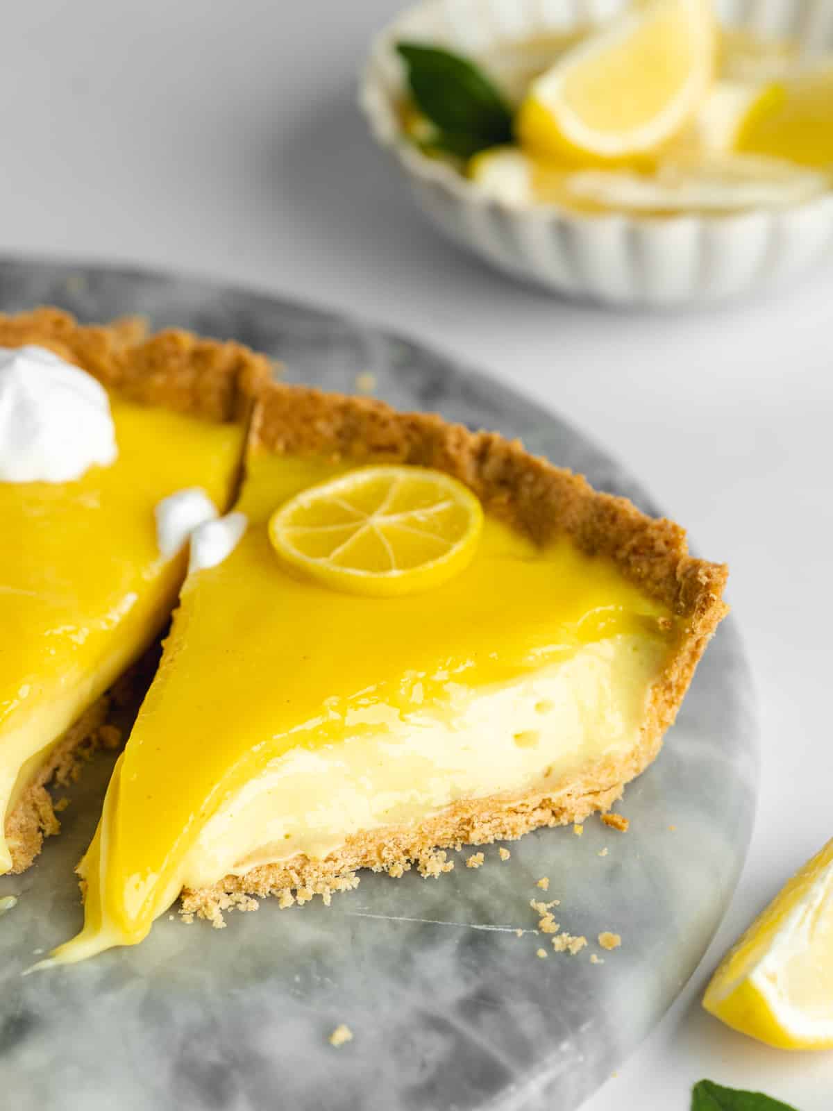 a slice of lemon curd with creamy filling and lemon curd topping and a small bowl of lemon wedges in the background.
