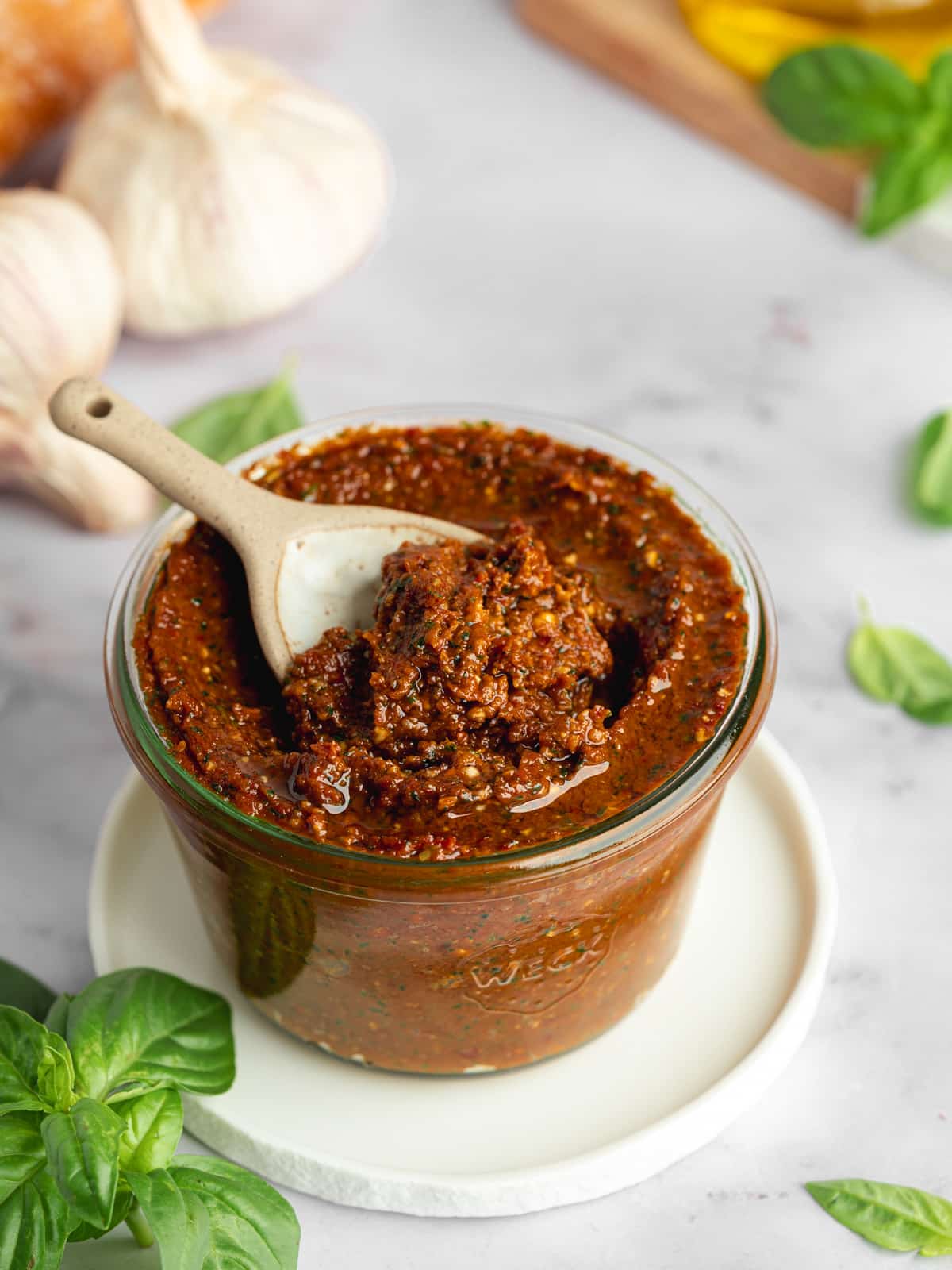 a jar of homemade red tomato pesto with a ceramic spoon inside and fresh basil leaves and garlic cloves scattered around it.