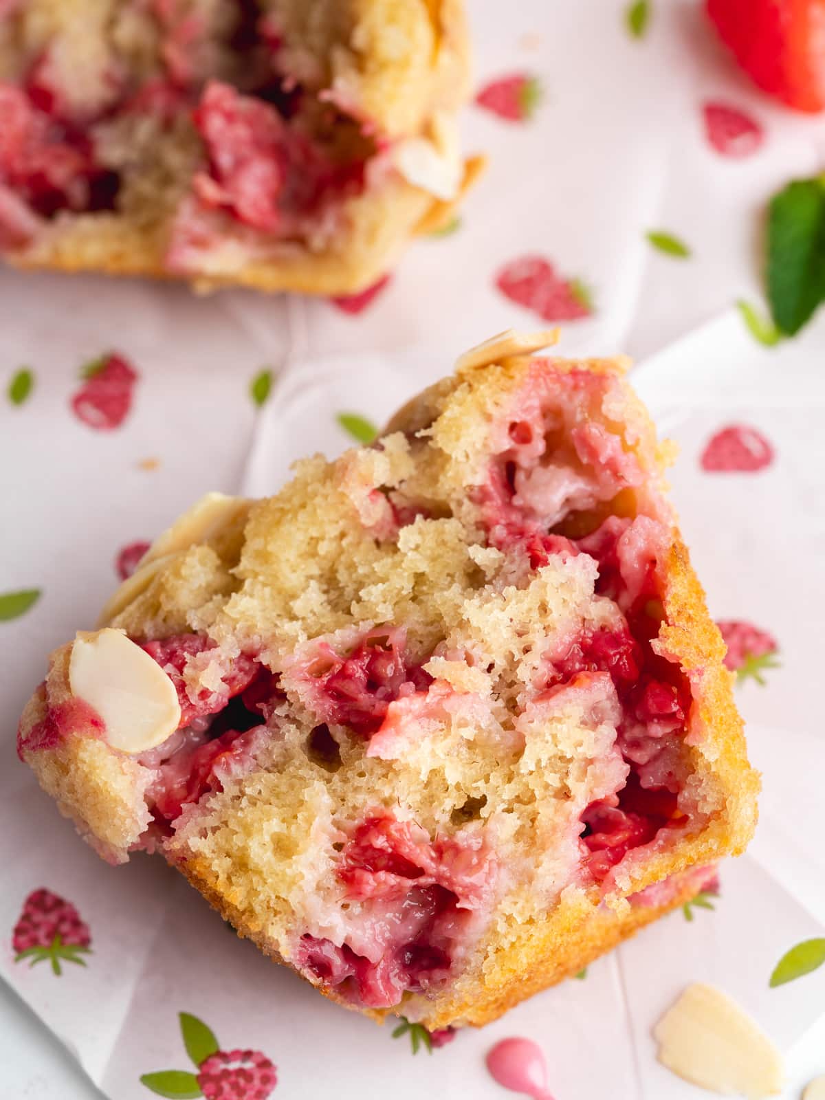 a muffin thats sliced in half and studded with frseh raspberries on raspberry-themed parchment paper.