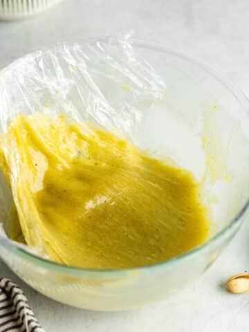 white chocolate pistachio ganache in a mixing bowl with plastic wrap being lifted away from the top.