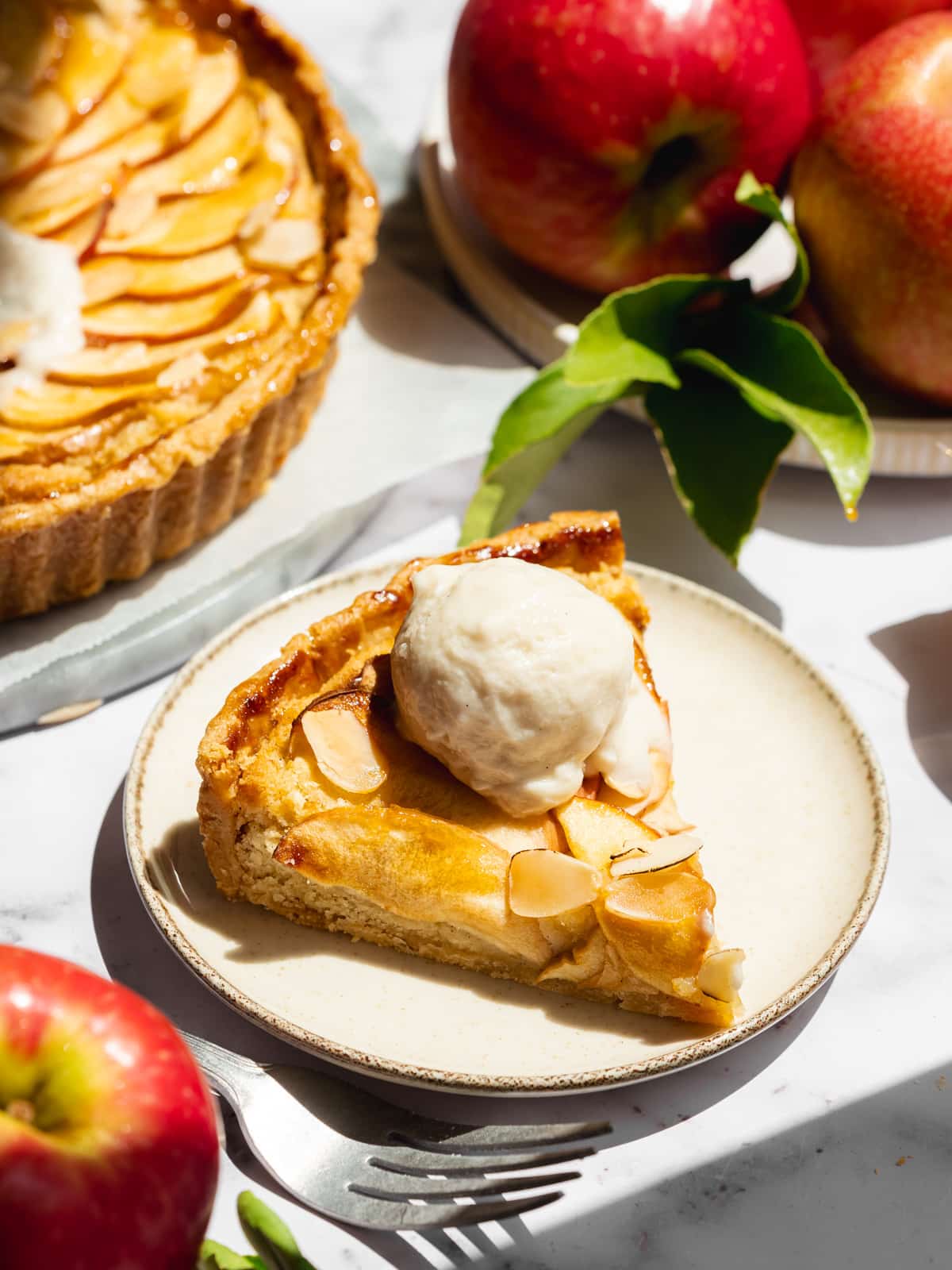 a slice of apple tart with a scoop of vegan ice cream on a ceramic plate with fresh red apples scattered around.