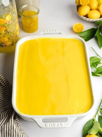 lemon tiramisu in a staub dish topped with a smooth layer of lemon curd with a bottle of limoncello on the side.