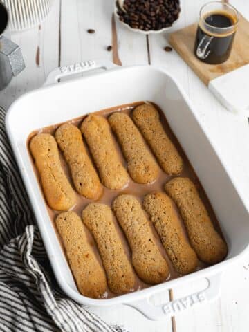a row of espresso-dipped ladyfingers in a white staub baking dish.