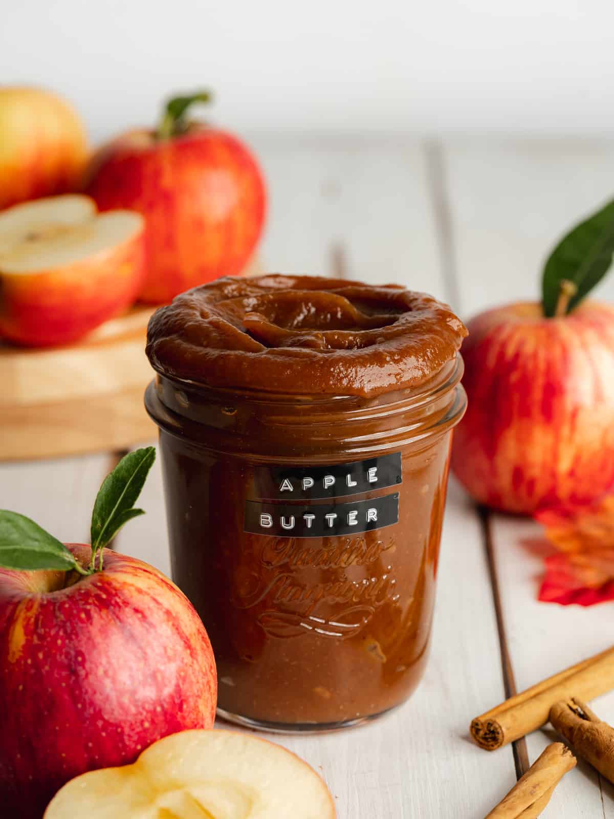 a jar of homemade apple butter with red apples and cinnamon sticks scattered around it.