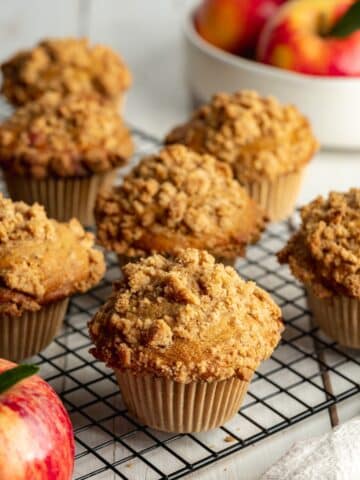 apple crumble muffins on cooling on a wire rack with a bowl of apples in the background.