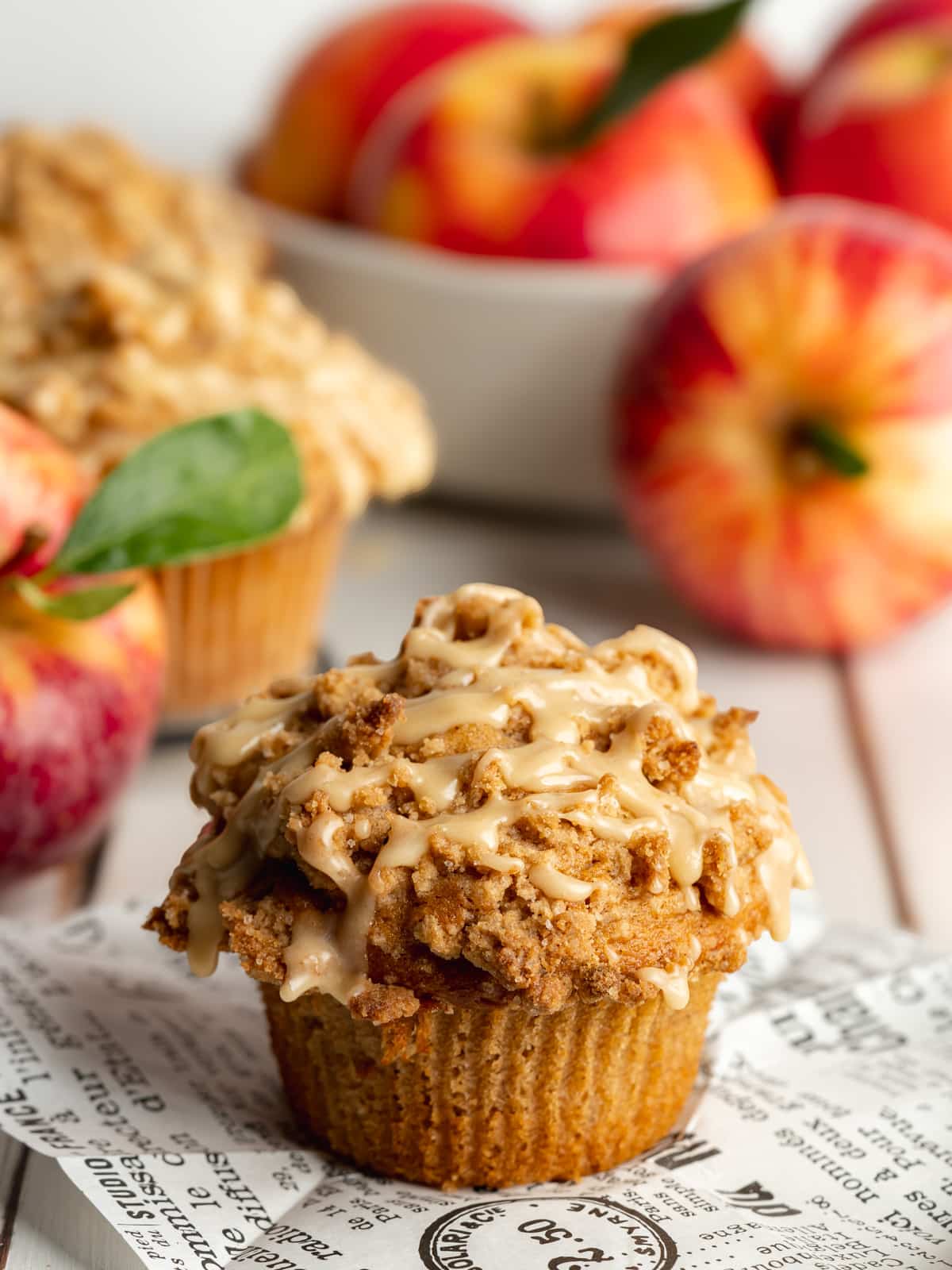 apple muffin with maple glaze on some newspaper with a bowl of fresh red apples in the background.