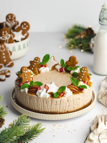 a gingerbread cheesecake decorated with gingerbread men, mint leaves, gingerbread stars and pomegranate, with cookies scattered around the scene.