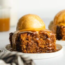 a slice of vegan sticky toffee pudding on a white plate with a scoop of ice cream on top and toffee sauce poured over it.