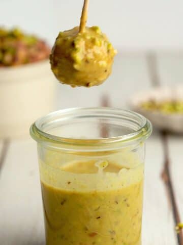 a pistachio truffle on a wooden skewer being dipped into pistachio melted white chocolate.