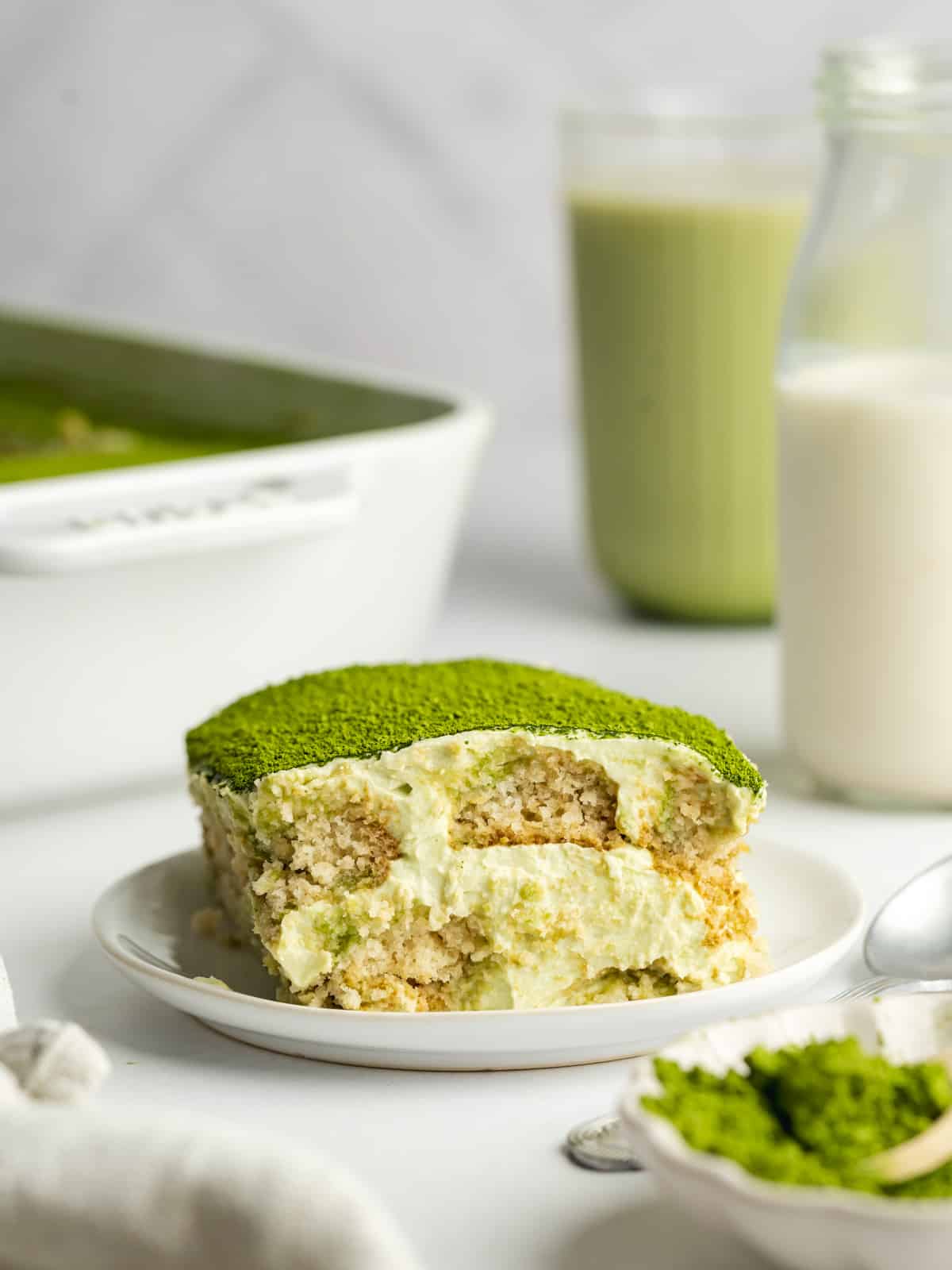 a slice of matcha tiramisu on a ceramic plate with a glass of matcha latte in the background.