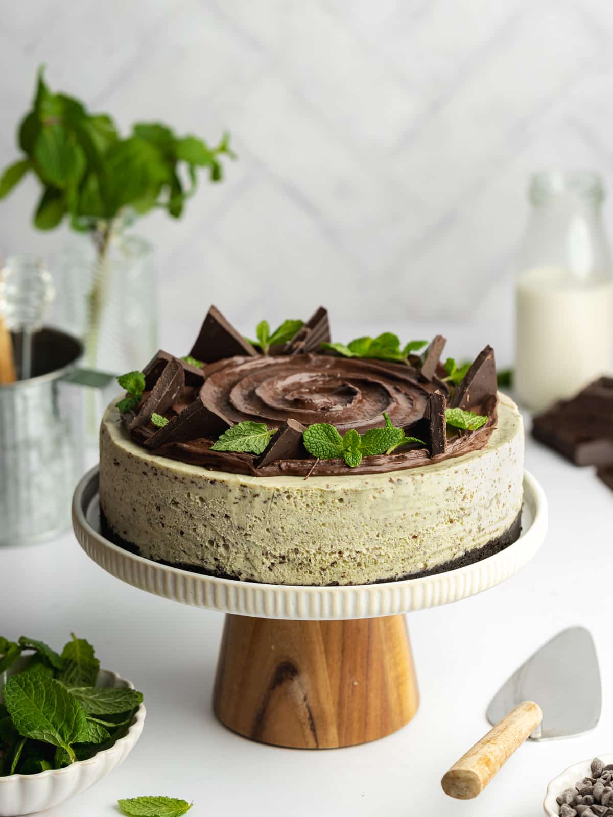 a mint chocolate chip cheesecake topped with ganache on a wooden cake stand.