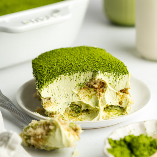 a slice of green tea tiramisu on a plate with matcha powder on top and a spoonful taken showing the creamy consistency.
