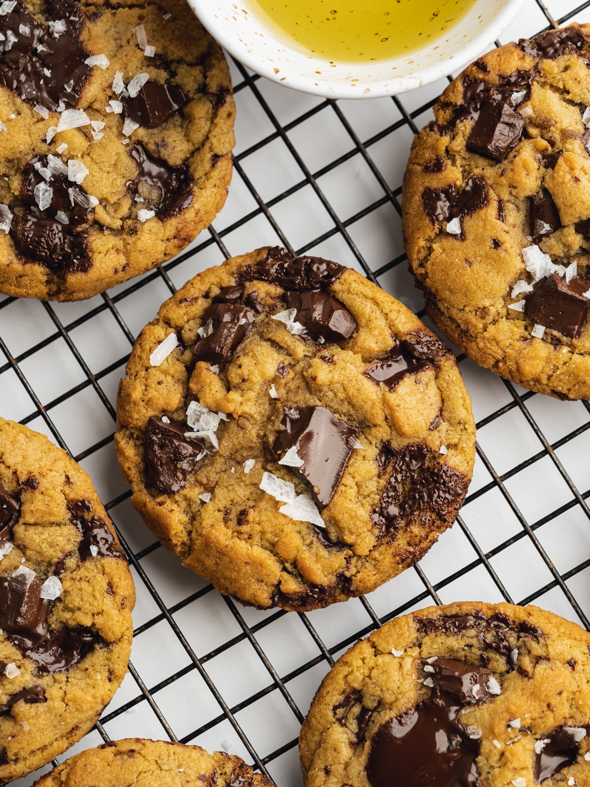 chocolate chunk olive oil cookies on a wire rack with flaky sea salt on top and a small pinch bowl with olive oil.