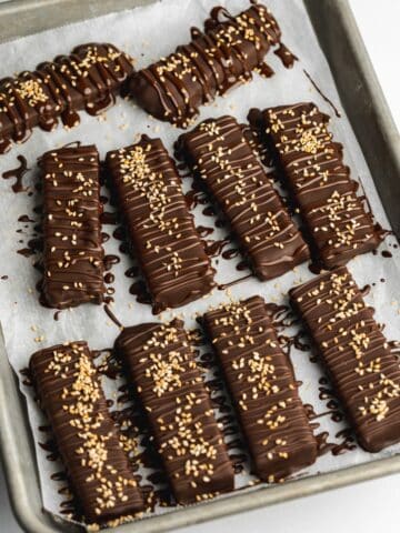 date bars coated in dark chocolate with sesame seeds on top of a tray lined with parchment paper.