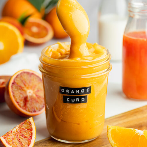a jar of orange curd with peeled oranges scattered around it.