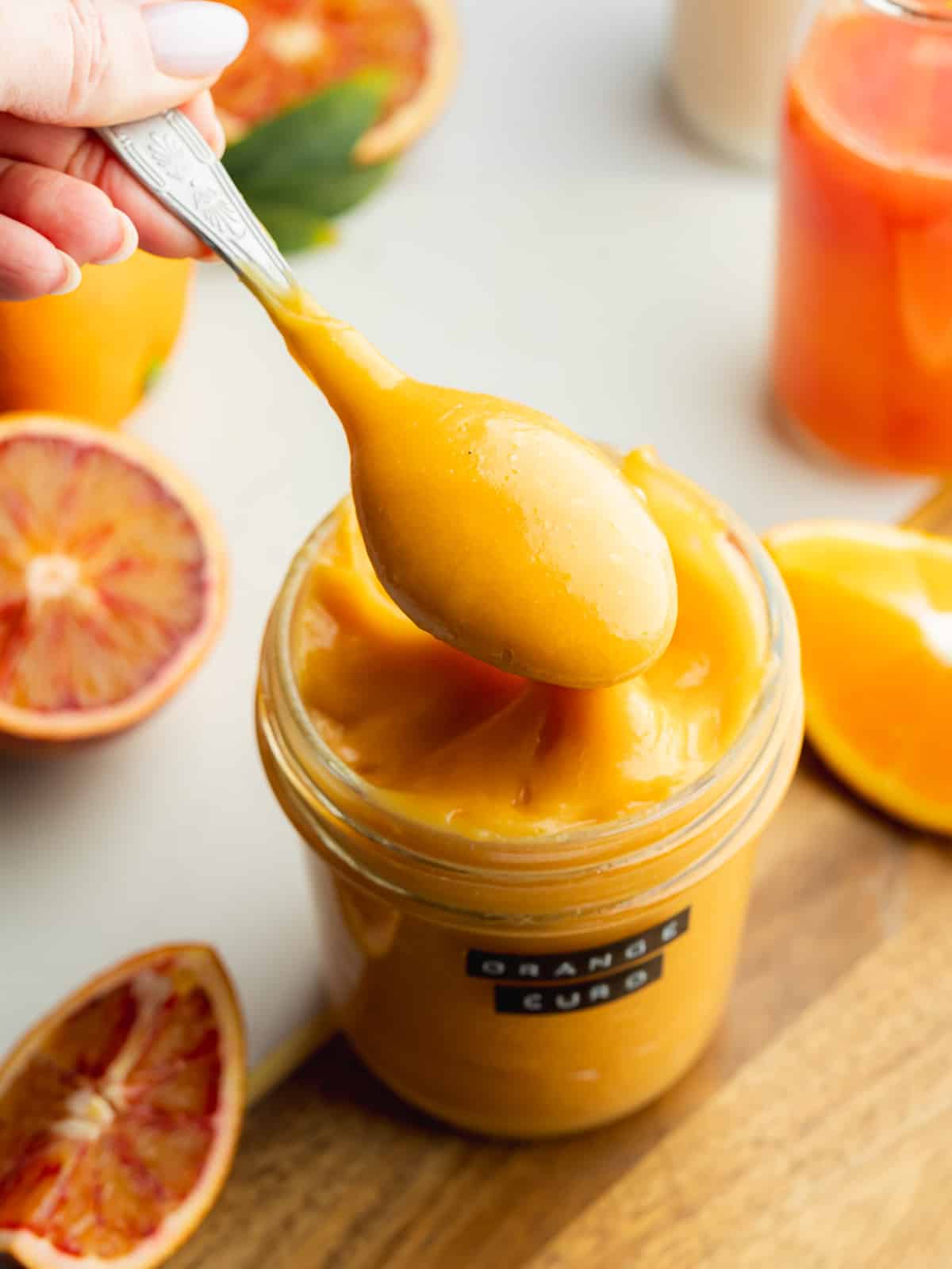 a jar of orange curd with a spoon showing the thick consistency and blood oranges scattered around it.