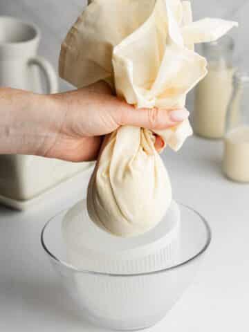 a hand holding up a cheesecloth with homemade macadamia nut cheese.