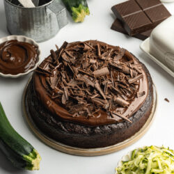 a chocolate zucchini cake topped with vegan ganache and chocolate curls with grated courgette and chocolate scattered around it.