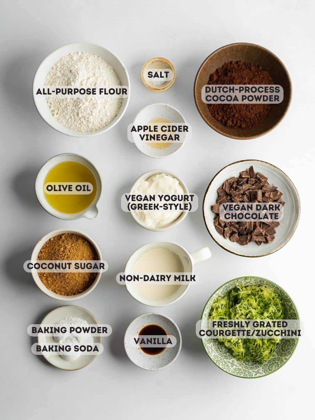 ingredients for chocolate zucchini muffins measured out in bowls on a light gray surface.
