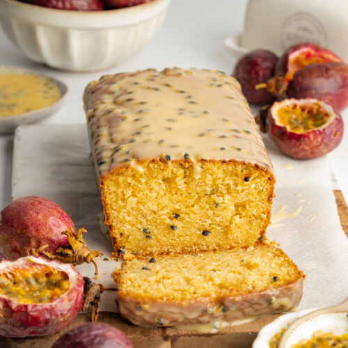 a passion fruit loaf cake on a cutting board with a skice cut from it showing the moist crumb, and fresh passion fruits scattered around it.