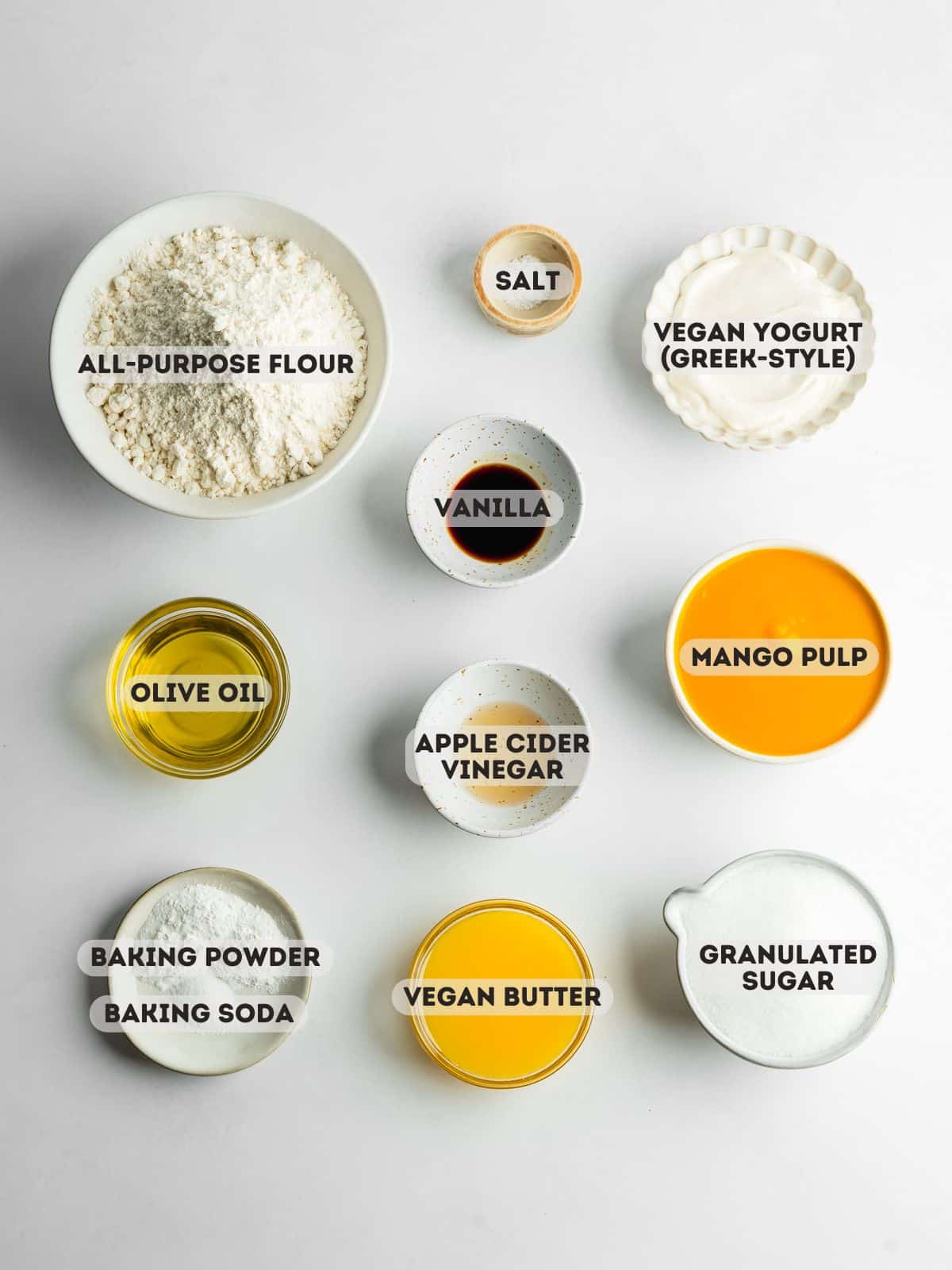 ingredients for mango cake measured out in bowls on a gray surface.