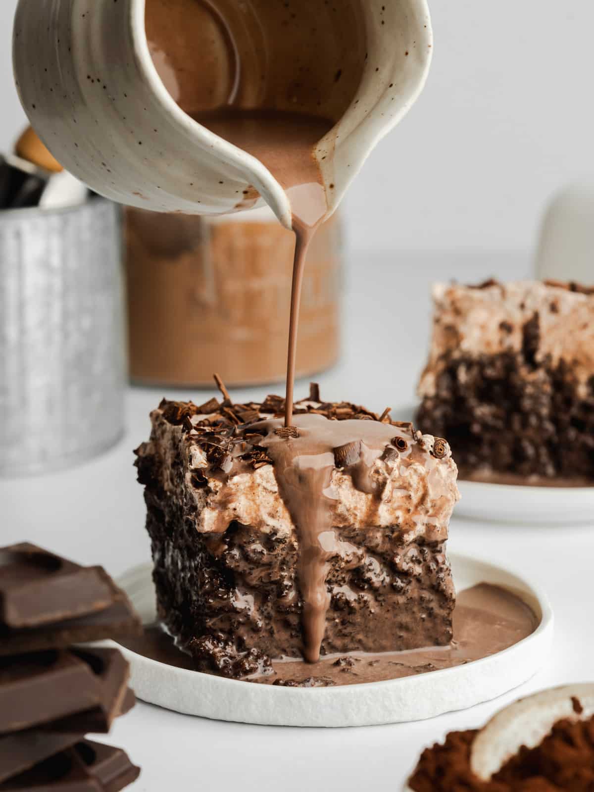 a slice of chocolate tres leches on a white ceramic plate with chocolate milk mixture being poured over the top.