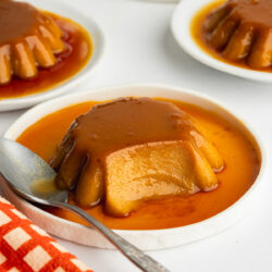 three mini pumpkin flans on small white plates with caramel sauce poured over them.
