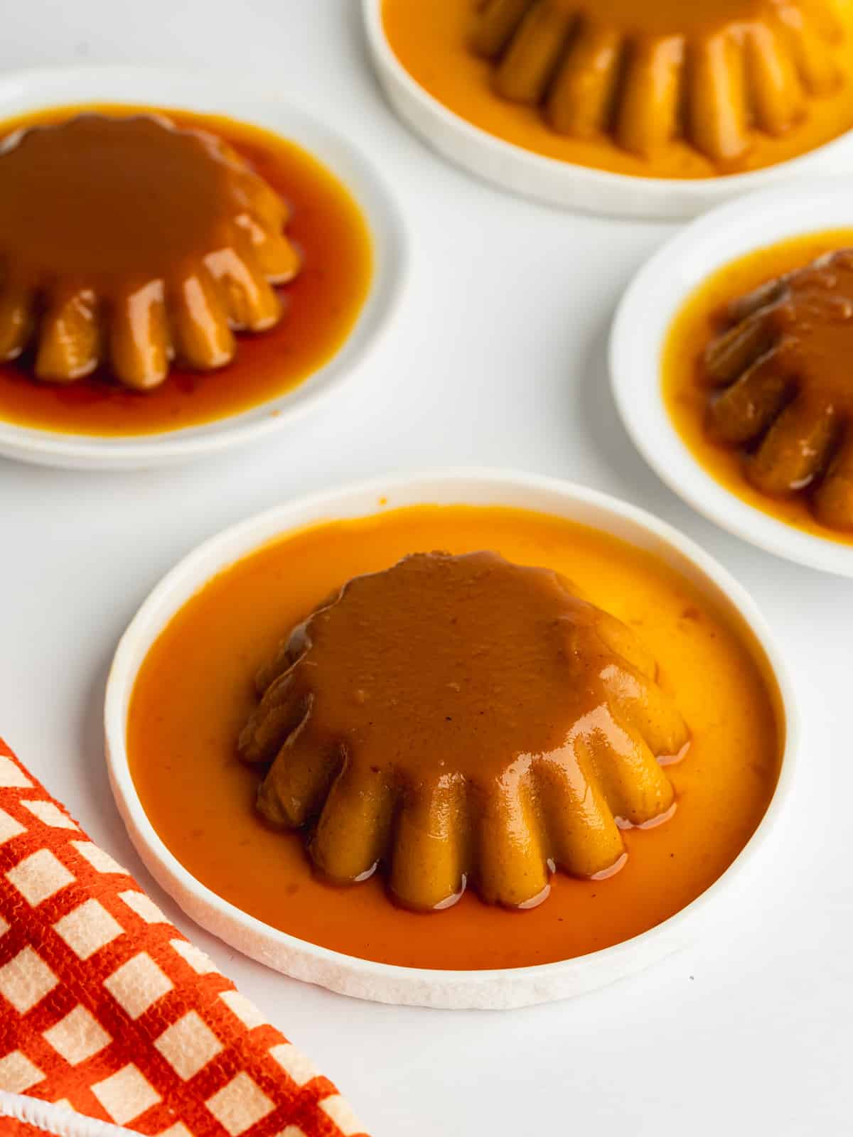 4 pumpkin flans unmolded on small white plates with runny caramel sauce cascading over the tops.