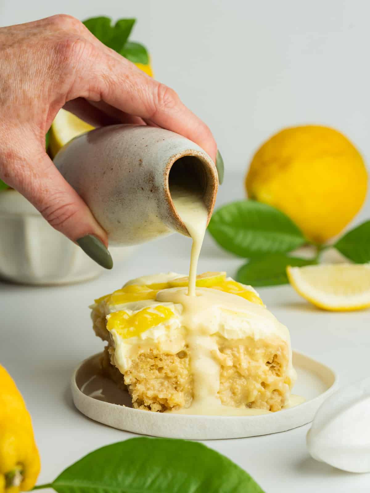 a slice of lemon tres leches on a ceramic plate with a jug of triple milk mixture being poured over the top.