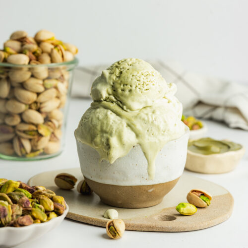 creamy scoops of pistachio ice cream in a ceramic ramekin with pistachios scattered around it.