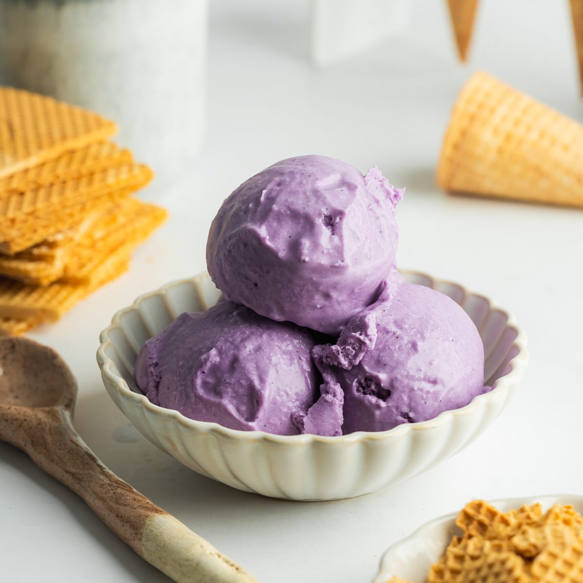 3 scoops of ube ice cream in a small ceramic bowl with ice cream cones and wafers in the background.