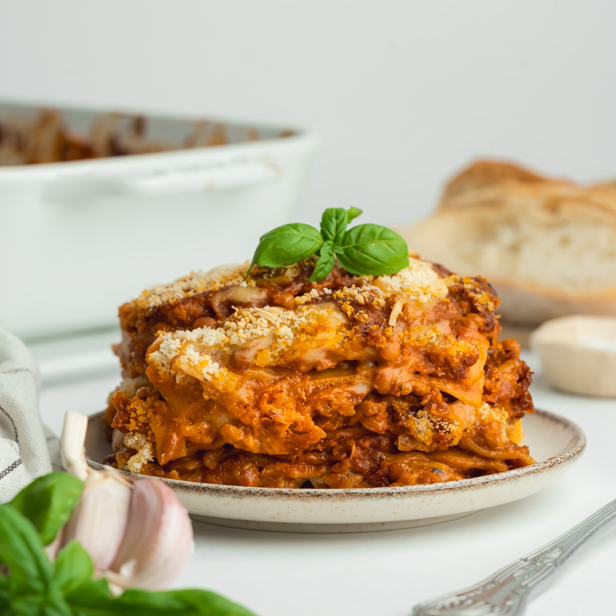 a slice of tall veegan lasagne on a white plate with fresh bread and a bottle of red wine in the background.