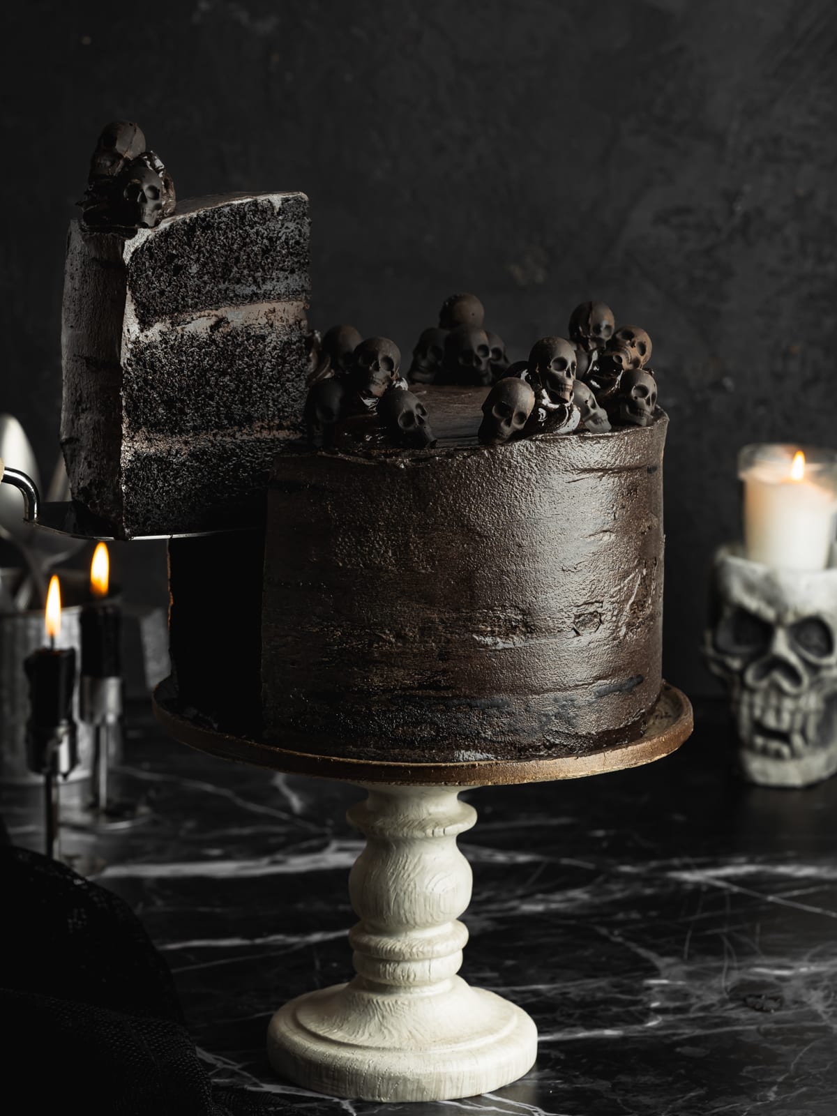 Vegan black velvet layer cake with a slice removed, revealing three layers of dark black cocoa sponge sandwiched with chocolate frosting and decorated with black skulls for Halloween.
