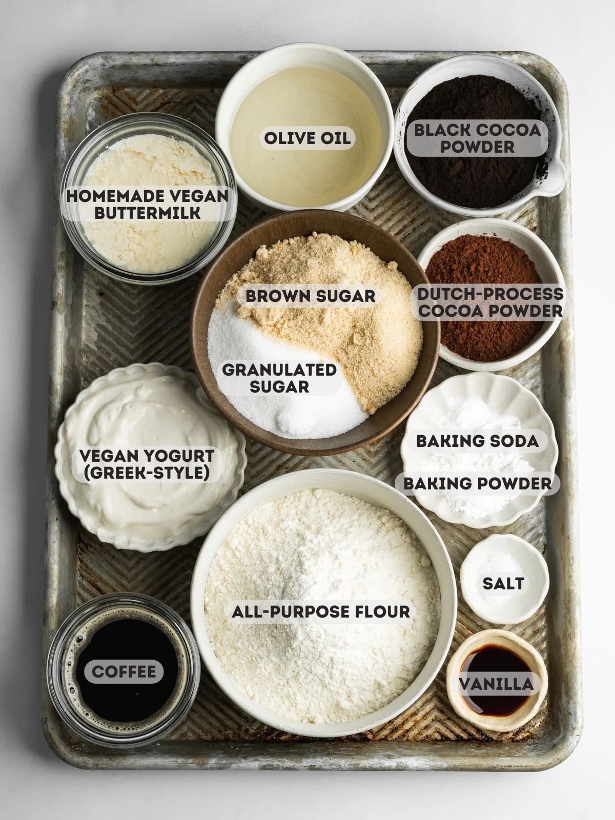 Overhead view of ingredients for vegan black velvet cake, including black cocoa powder, Dutch-process cocoa, flour, sugar, olive oil, vegan buttermilk, yogurt, coffee, vanilla, and raising agents.