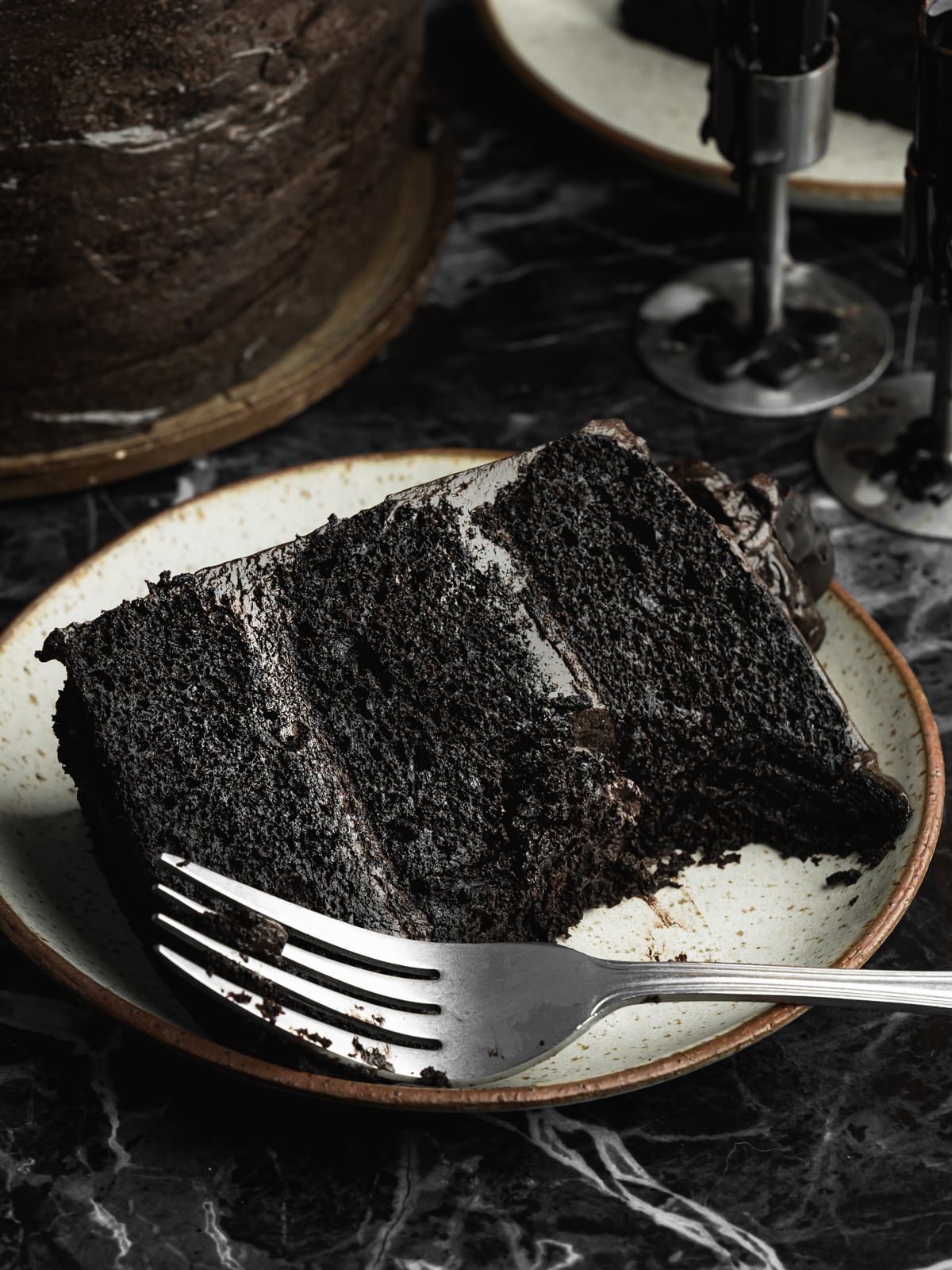Slice of vegan black velvet cake with three layers of moist black cocoa sponge and rich chocolate frosting, served on a ceramic plate with a silver fork.