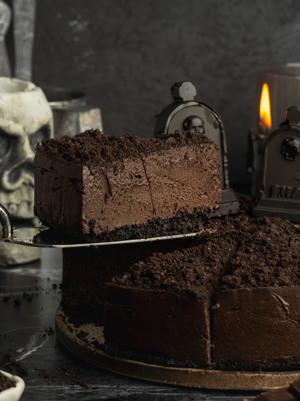 Slice of vegan dark chocolate Halloween cheesecake with Oreo crust, chocolate cookie crumbs, and tombstone decorations.