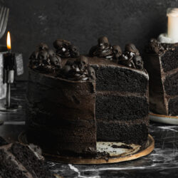Vegan black cocoa Halloween cake covered in dark chocolate frosting and decorated with miniature black skulls, displayed on a vintage cake stand surrounded by candles.